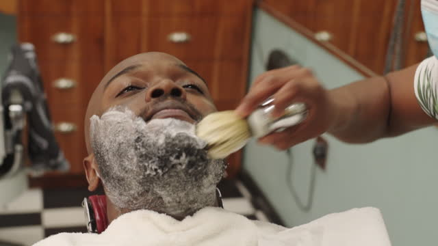 Close-up of a barber applying shaving cream with a brush to the beard of a client lying in a chair in his barbershop