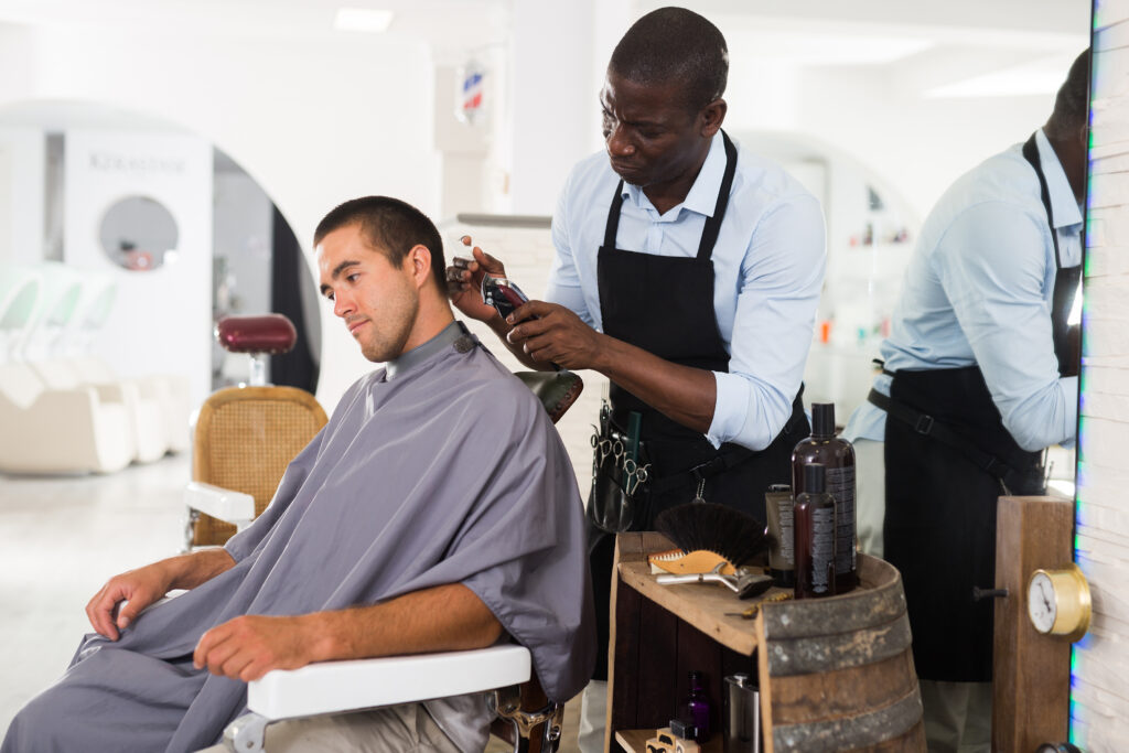 African-American hairdresser doing styling for young man with hair clipper at barber shop
