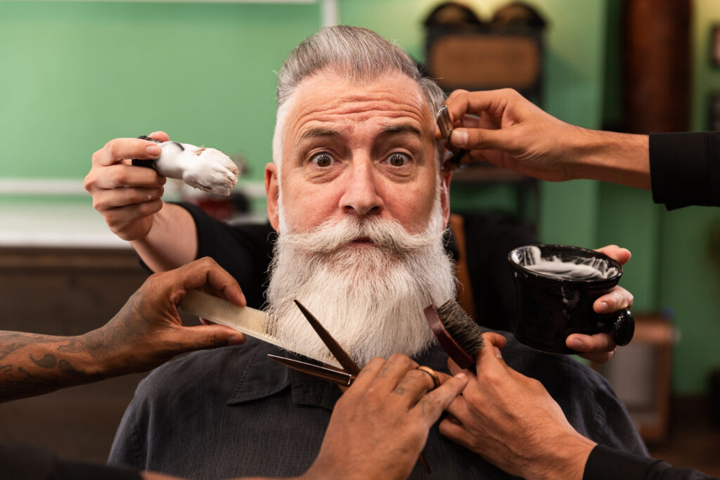 mature man with white beard looking at camera in barber shop with barber hands with cutting and shaving instruments, brush, scissors, comb, razor. hipster style